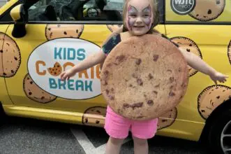 A small child dressed as a cookie behind a car designed around the company :"Kids Cookie Break"